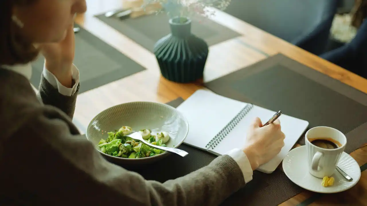 A small business owner sits at a table with a notebook, coffee, and a healthy meal, reflecting on financial decisions. This image represents how everyday routines and mindful choices support better money habits for small business owners.